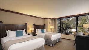 Een hotelkamer met twee bedden, een bureau en een groot raam met uitzicht op bomen en een besneeerd landschap.