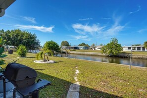 A grassy area with a water body, a barbecue grill, and a pathway leading to a building. - Waterfront Gulf Access Beach House w Lainai (Port Charlotte)