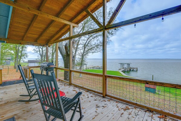 A covered deck with a view of a lake, wooden chairs, a grill, and a dock. - Dock + Fire Pit: Tranquil Home on Lake Livingston (Livingston)