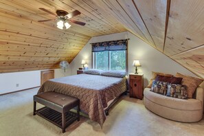 Une chambre avec un plafond en pente, un lit recouvert d’une courtepointe, un banc, une table de chevet avec une lampe et une fenêtre donnant sur des arbres.