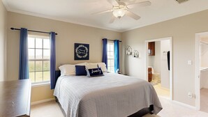 Una camera da letto con un letto, una ventola a soffitto, una finestra con tende, un elemento decorativo a parete e una porta che conduce a un'altra stanza.