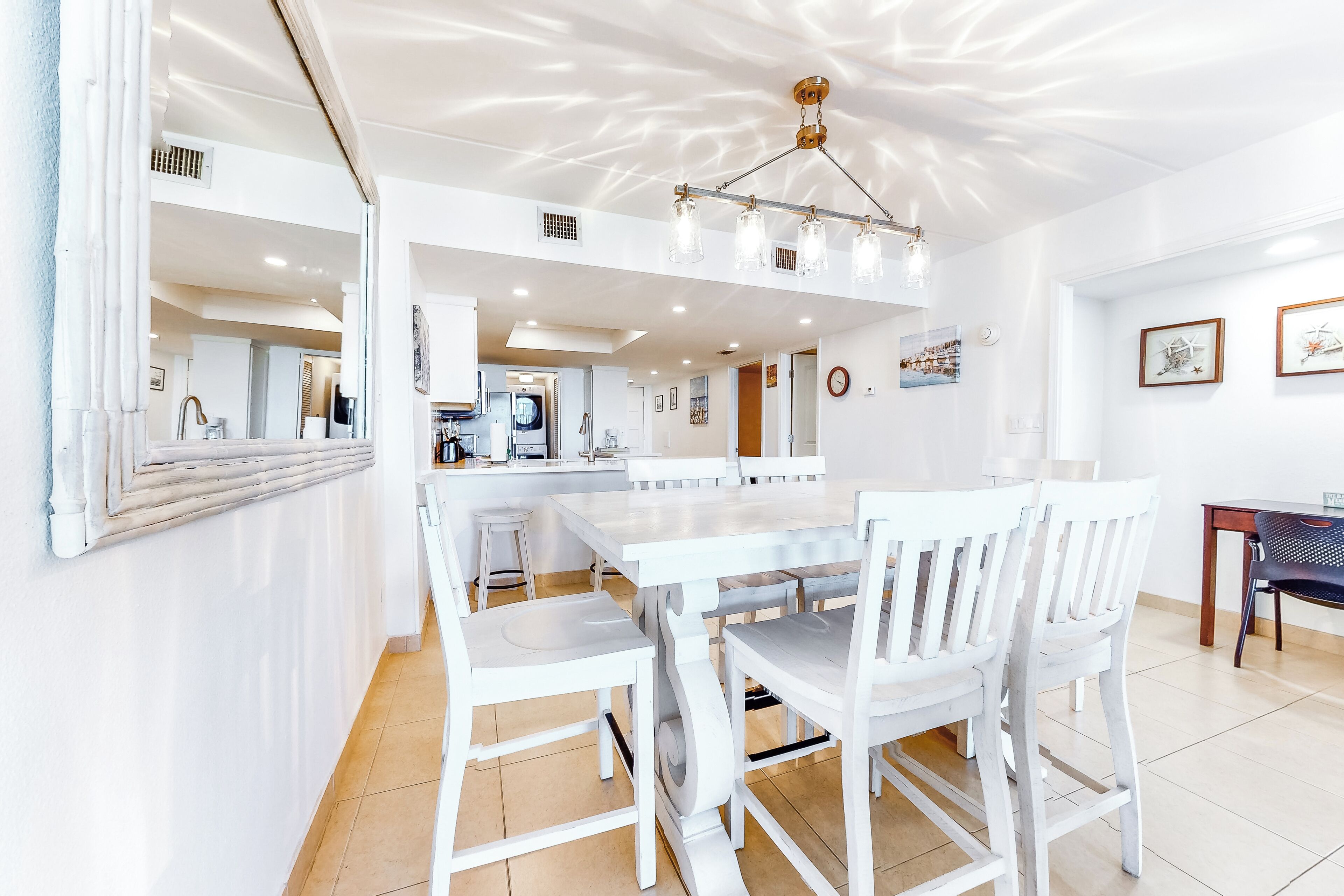 A spacious dining area with a white table and chairs, a kitchen in the background, and a ceiling light fixture.