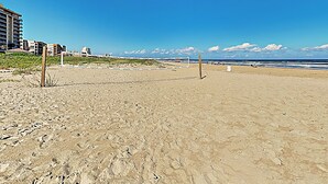 A beach with a volleyball net, sandy terrain, and buildings in the background. - 2BR w/ Gulf-view Balcony: Suntide III - Unit 307 (South Padre Island)