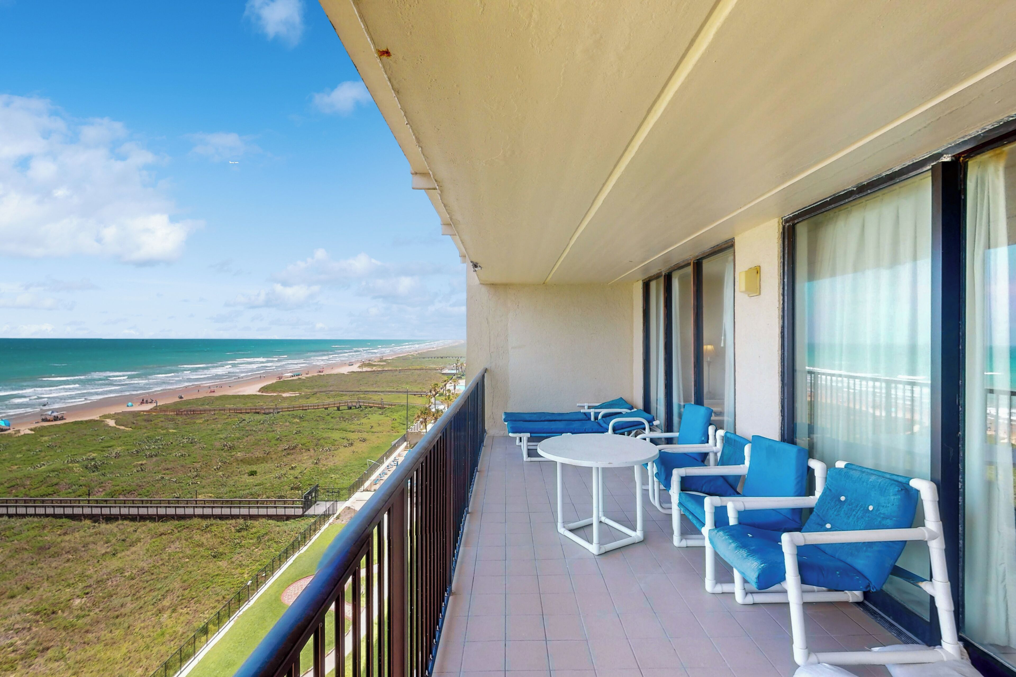 Un balcon avec des chaises bleues et une petite table, donnant sur une plage et l’océan.