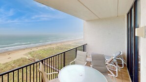 Un balcón con una mesa y sillas de vidrio, con vistas a una playa y una vista despejada del océano.