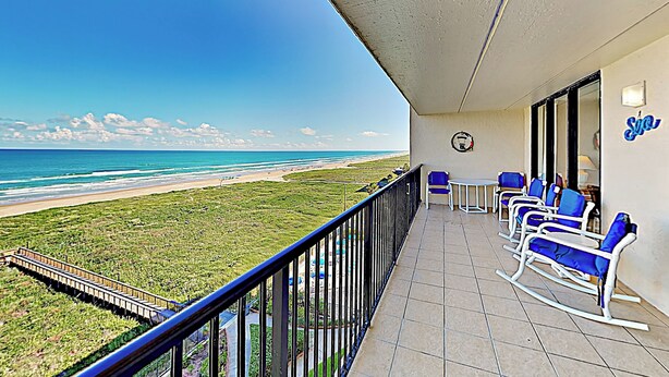 A balcony with a view of the beach, a table, and blue chairs. - 3BR Gulf-view Suite w/ Pools, Gym & Tennis (South Padre Island)