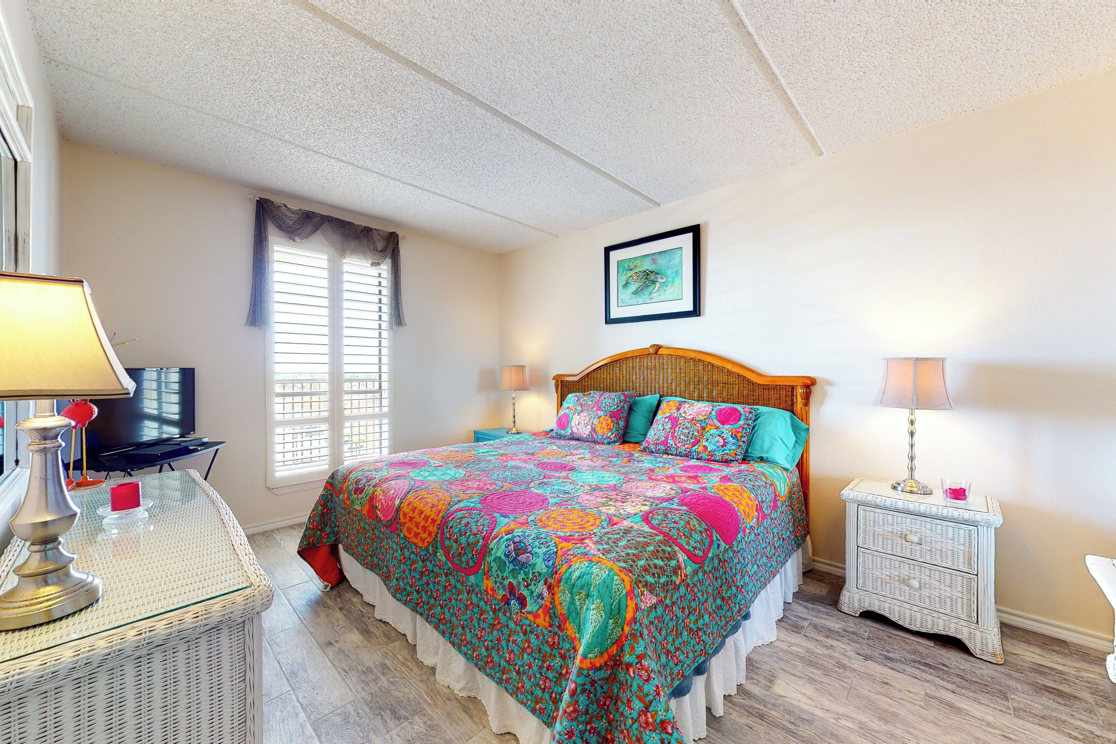 A bedroom with a large bed, a television, a desk with a lamp, and a wicker nightstand.