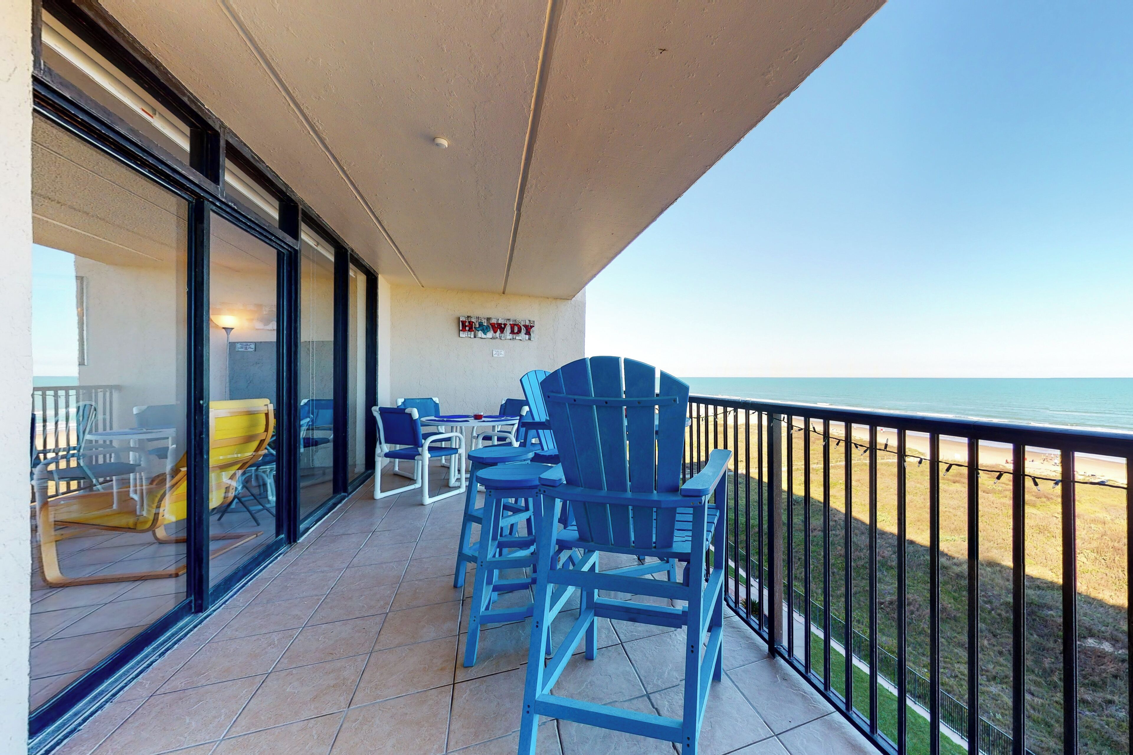 Un balcón con sillas Adirondack azules, una mesa y sillas, con vista a una playa.