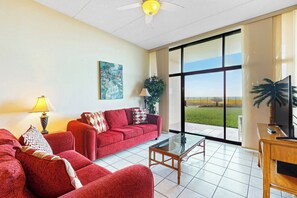 A spacious living room with a red sofa set, a glass coffee table, and a large window offering a view of the outdoors. - Resort 2BR w/ Patio, Perks: Suntide III - Unit 104 (South Padre Island)