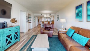 Ein geräumiges Wohnzimmer mit einem braunen Ledersofa, einem Holztisch und einem blaugrünen Teppich. Im Hintergrund befindet sich ein Essbereich mit Tisch und Stühlen.