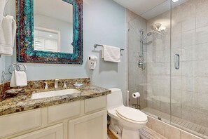 A bathroom with a granite countertop sink, a white toilet, a glass-enclosed shower, and a decorative mirror with a colorful frame. - 2BR Penthouse Beach Perch: Suntide III - Unit 1207 (South Padre Island)
