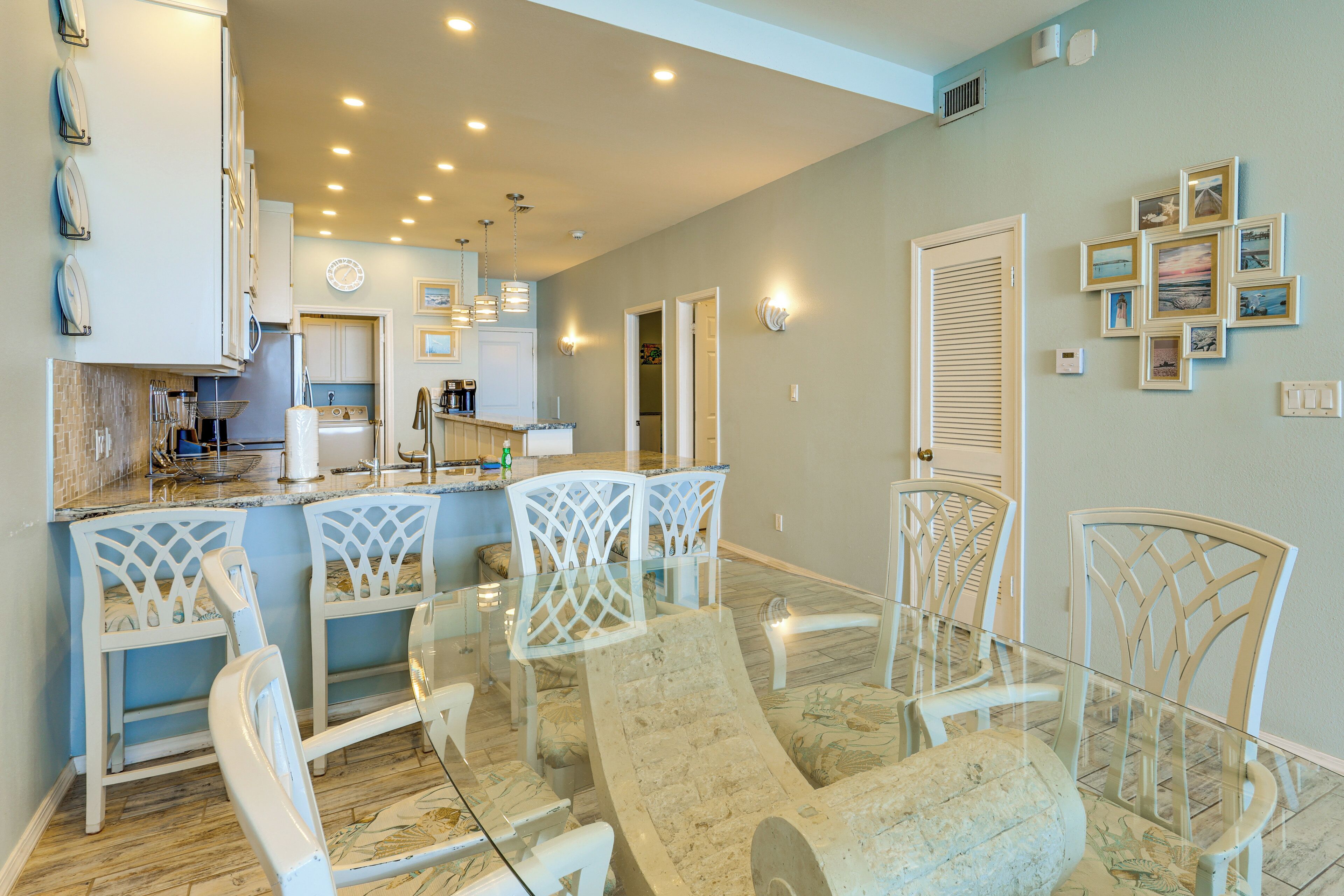 A modern kitchen with white cabinetry, a dining area with white chairs and a glass table, and a hallway with a door and wall-mounted pictures.
