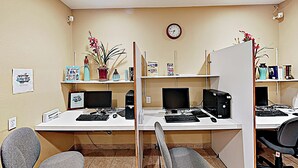 Et professionelt kontormiljø med flere arbejdsstationer, hver med en computer og et tastatur. Der er plantet potteplanter på hylderne, og et ur er synligt på væggen.