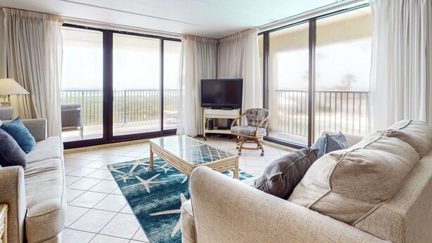 Ein geräumiges Wohnzimmer mit einer Couch, einem Sessel und einem Glastisch. Ein Flachbildfernseher ist an der Wand befestigt, und es gibt einen Balkon mit Blick.