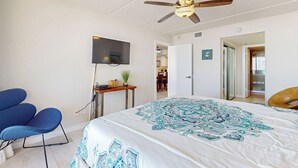 Une chambre à coucher avec un lit, un ventilateur de plafond, une télévision, un bureau avec une plante et une chaise.