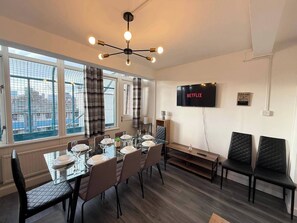 A modern dining area with a glass table set for six, a TV mounted on the wall, and a city view through large windows. - Modern Bright Top-floor Flat (London)