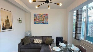A modern living room with a dark sofa, a glass dining table set for four, and a ceiling light fixture. - Modern Bright Top-floor Flat (London)