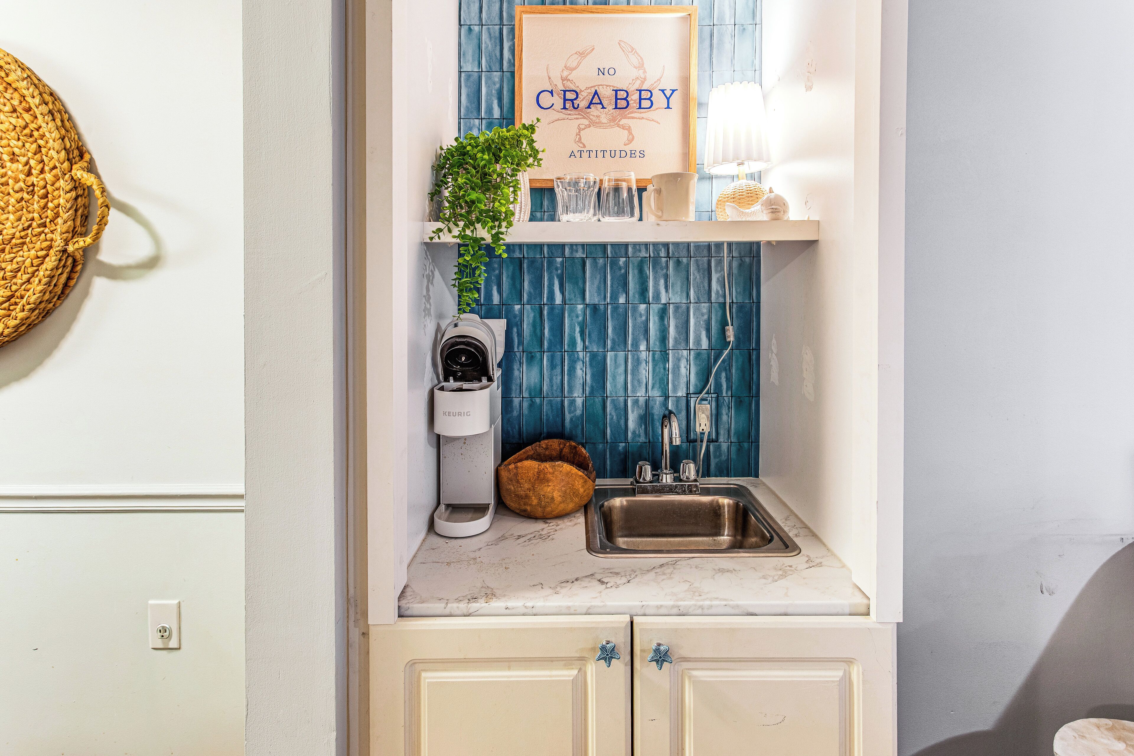 A kitchen with a sink, a coffee maker, a plant, and a framed sign on the wall.