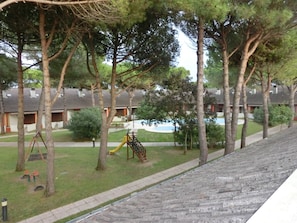 Un cortile con un'area giochi, alberi e una fila di edifici, sullo sfondo si intravede una piscina.