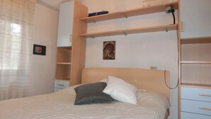 A bedroom with a bed, a bedside table, a nightstand, a wall-mounted shelf with a picture, and a window with curtains. - Cozy Apartment With Swimming Pool Access (San Michele al Tagliamento)