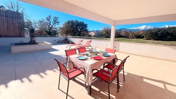 Eine Terrasse im Freien mit Tisch für vier Personen, umgeben von roten Stühlen, mit Blick auf eine reizvolle Landschaft aus Bergen und Bäumen.