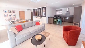 Ein modernes Wohnzimmer mit einem grauen Sofa, einem roten Sessel und einem kleinen runden Tisch. Im Hintergrund ist ein Küchenbereich mit Esstisch und Barhocker zu sehen.