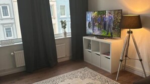 A living room with a television on a stand, a floor lamp, a patterned rug, and a window with curtains. - Vacation apartment Hansetraum Lübeck (Lübeck)