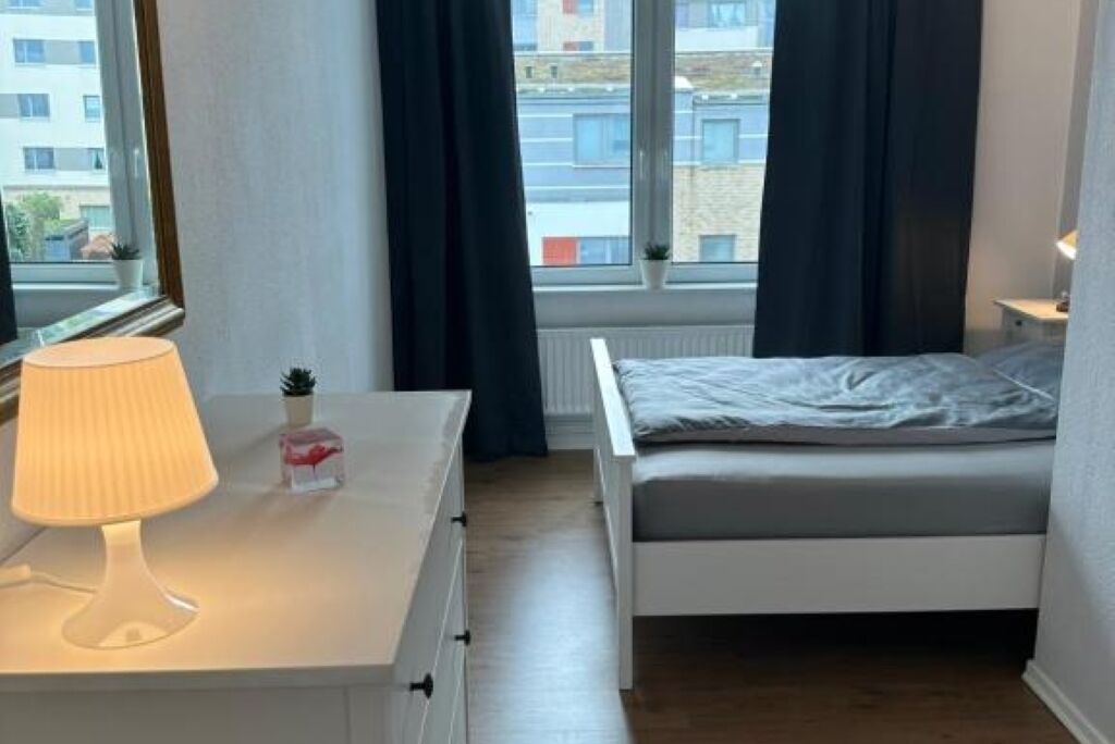 Un dormitorio con cama, una mesita de noche con lámpara, y vistas a edificios a través de la ventana.