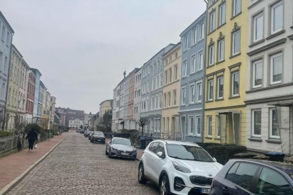 Eine Kopfsteinpflasterstraße gesäumt von bunten Gebäuden, geparkten Autos und einem Fußgänger.