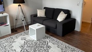 A modern living room with a dark grey sofa, white cushions, a small white side table, a floor lamp, and a patterned rug. - Vacation apartment Hansetraum Lübeck (Lübeck)