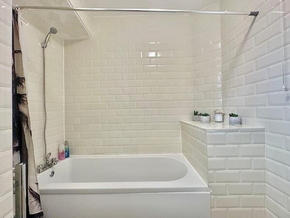 A bathroom with white tiled walls, a bathtub, a showerhead, and a shelf with toiletries. - Visit and Stay at Whitechapel (London)