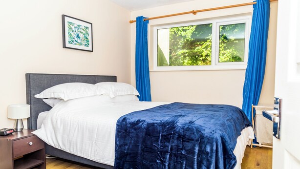 A neatly made bed with a white comforter and a blue blanket, a bedside table with a lamp, a framed picture on the wall, and a window with blue curtains.