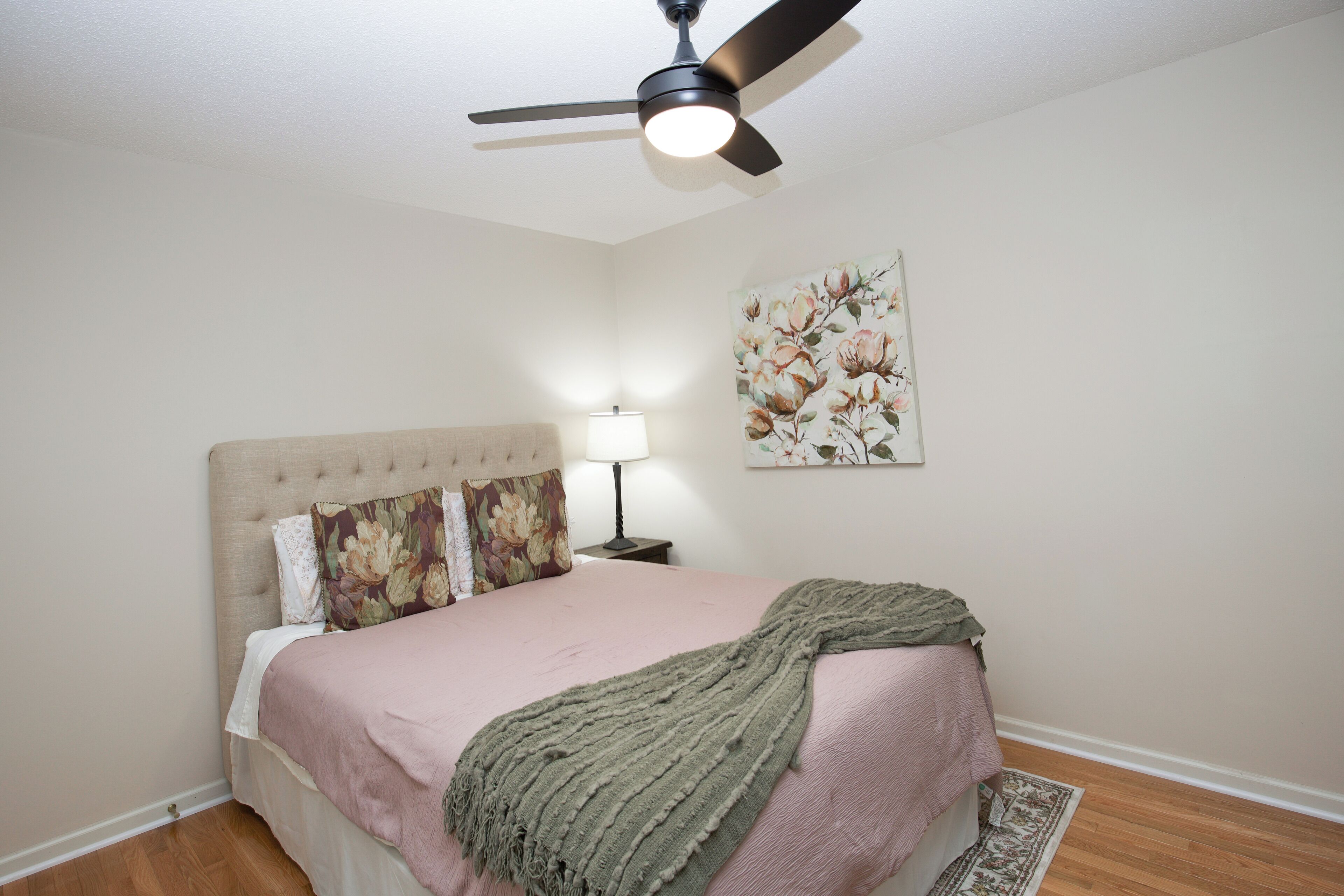 A bedroom with a bed, a ceiling fan, a nightstand with a lamp, and a floral painting on the wall.