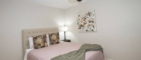 A bedroom with a bed, a ceiling fan, a nightstand with a lamp, and a floral painting on the wall.