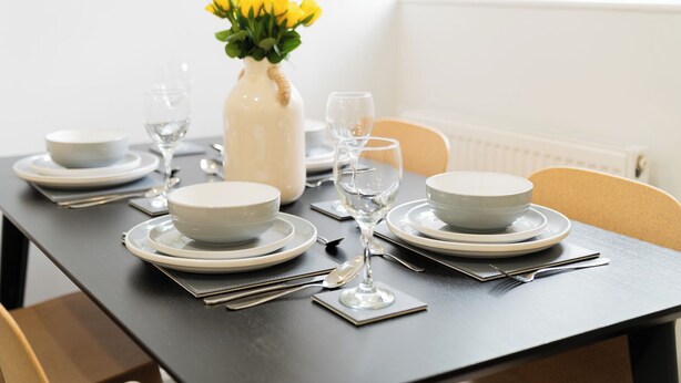Una mesa de comedor dispuesta con platos, cuencos y cubiertos blancos, acompañada de un florero con flores amarillas y una vela.