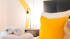 Una cama bien hecha con cabecera gris, almohada amarilla y una pila de toallas blancas.
