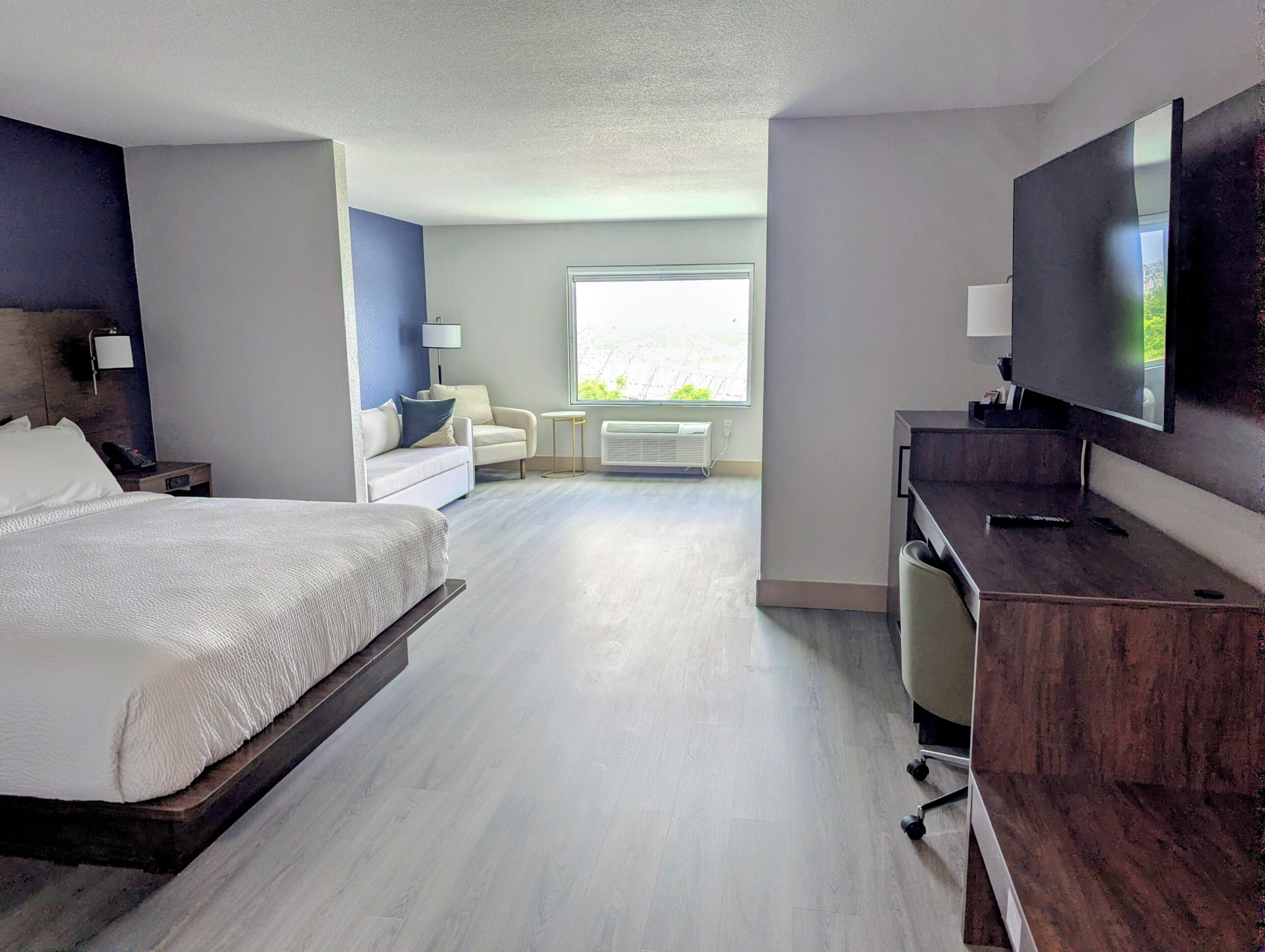 A hotel room with a bed, a desk, a chair, a television, a window, and a lamp.