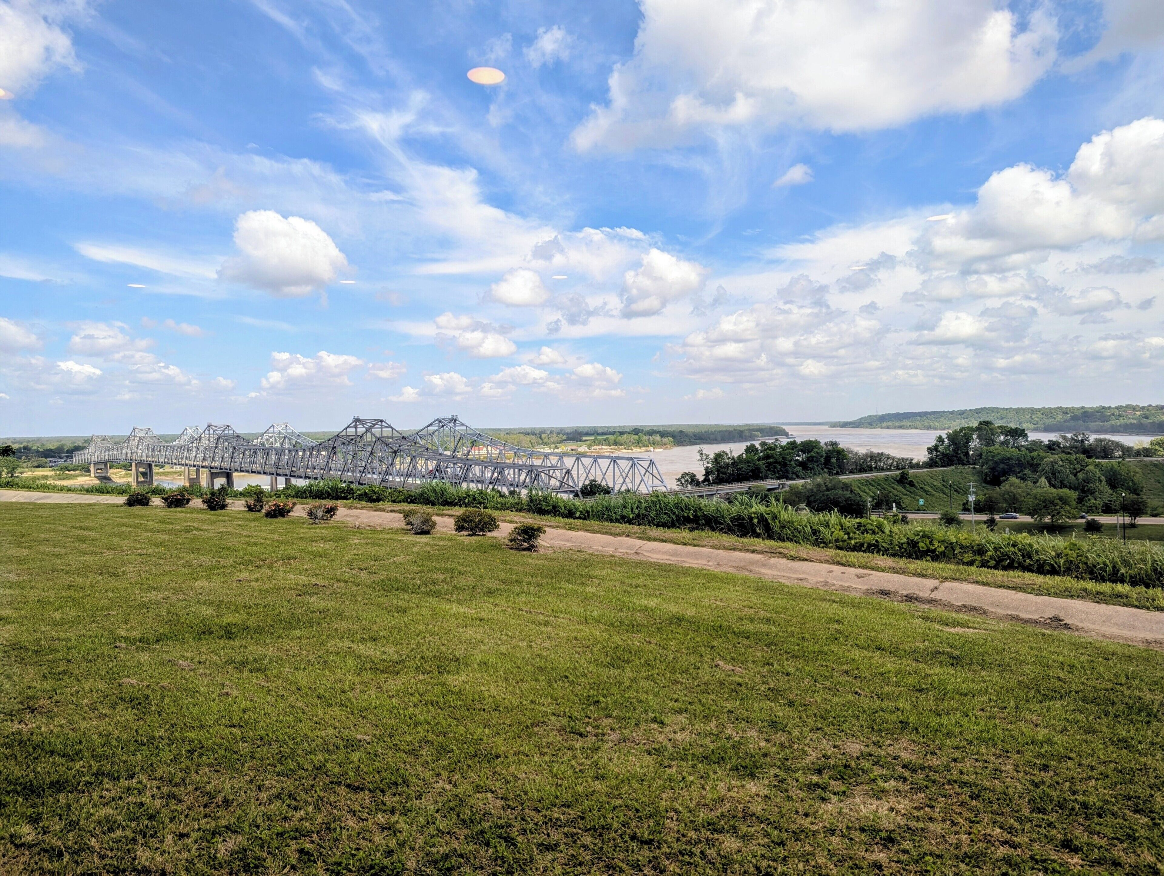 Eine große Metallbrücke überspannt einen Fluss, umgeben von grünen Feldern und Bäumen.