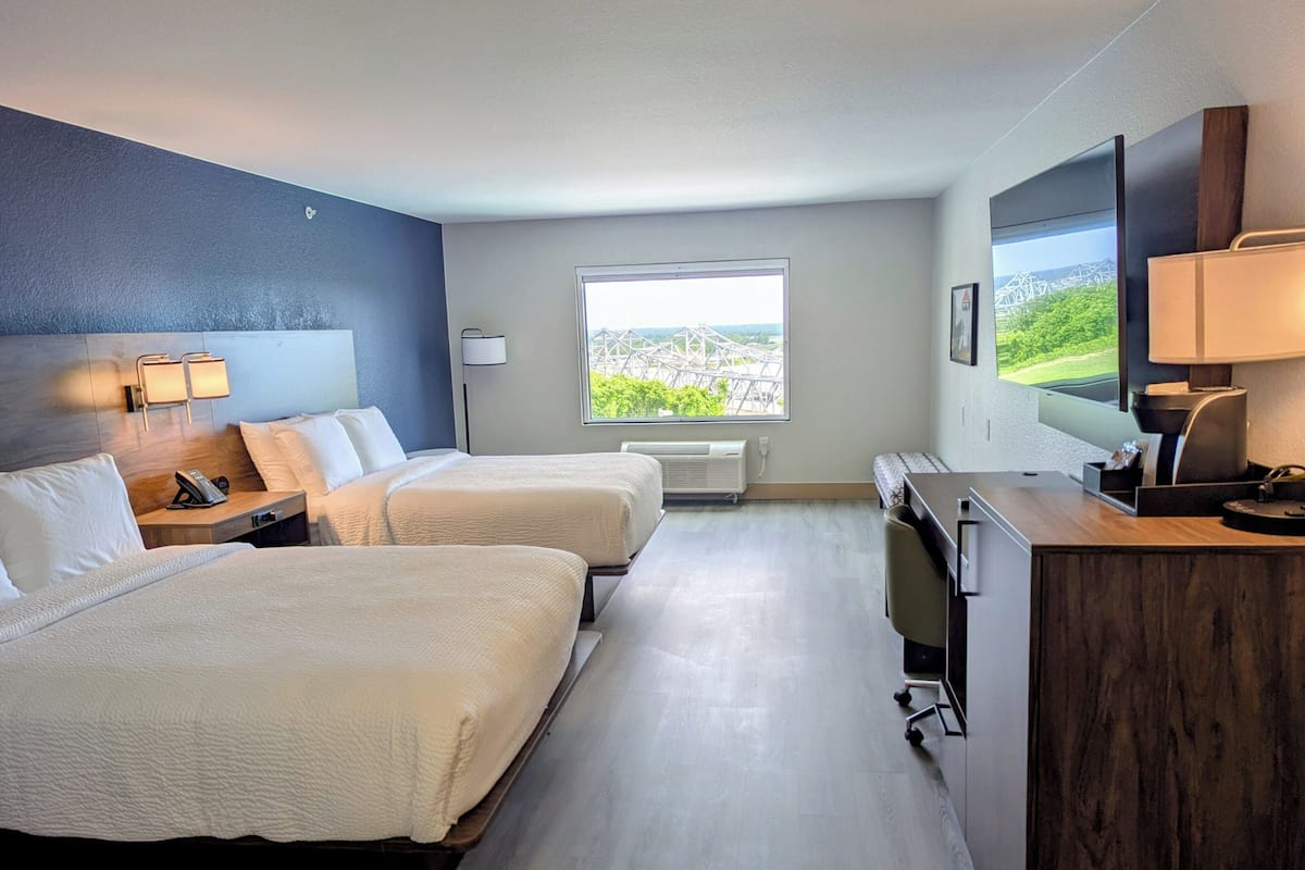 Habitación de hotel con dos camas, un escritorio, un televisor y ventana con vista a un puente.
