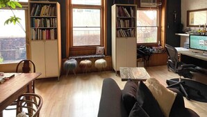 A cozy living room with a wooden floor, a black sofa, a dining table with chairs, and two bookshelves filled with books. There are three small stools and a decorative lamp. - Artist Loft in the Heart of Chelsea with BBQ & terrace, Pet friendly! (New York)