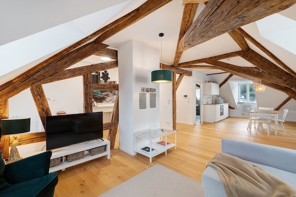 Ein Dachzimmer mit freiliegenden Holzbalken, einem Flachbildfernseher, einer Sitzecke, einem Essbereich und einer Küche.