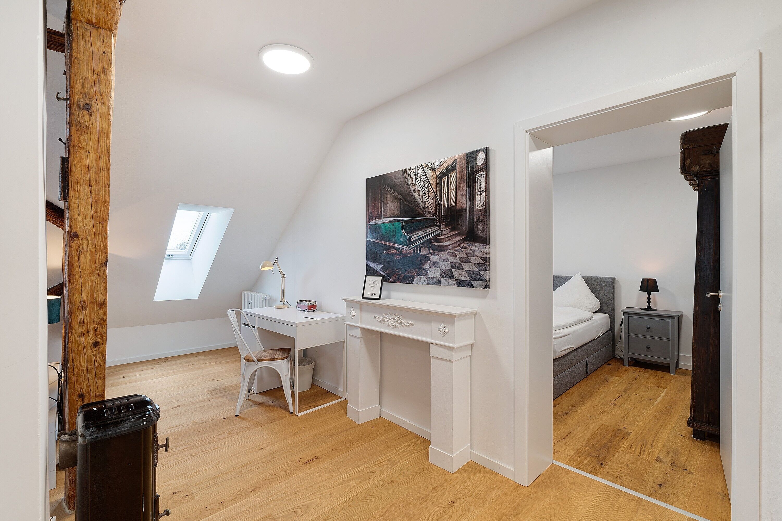 A bedroom with a bed, a desk, a chair, a nightstand, and a painting.