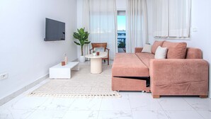 Un salon moderne avec un canapé, une table basse, un téléviseur fixé au mur et une vue sur l’extérieur grâce à de grandes fenêtres.