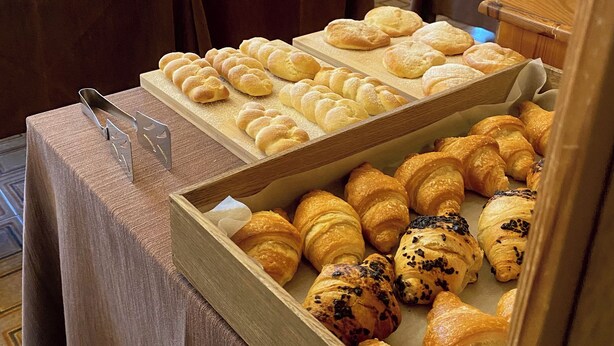 Een bakkerijdisplay met diverse gebakjes en brood, houten tafels en uitzicht op het buitengebeuren door grote ramen.