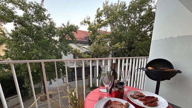 A balcony with a red table, two plates of food, a grill, and a potted plant.