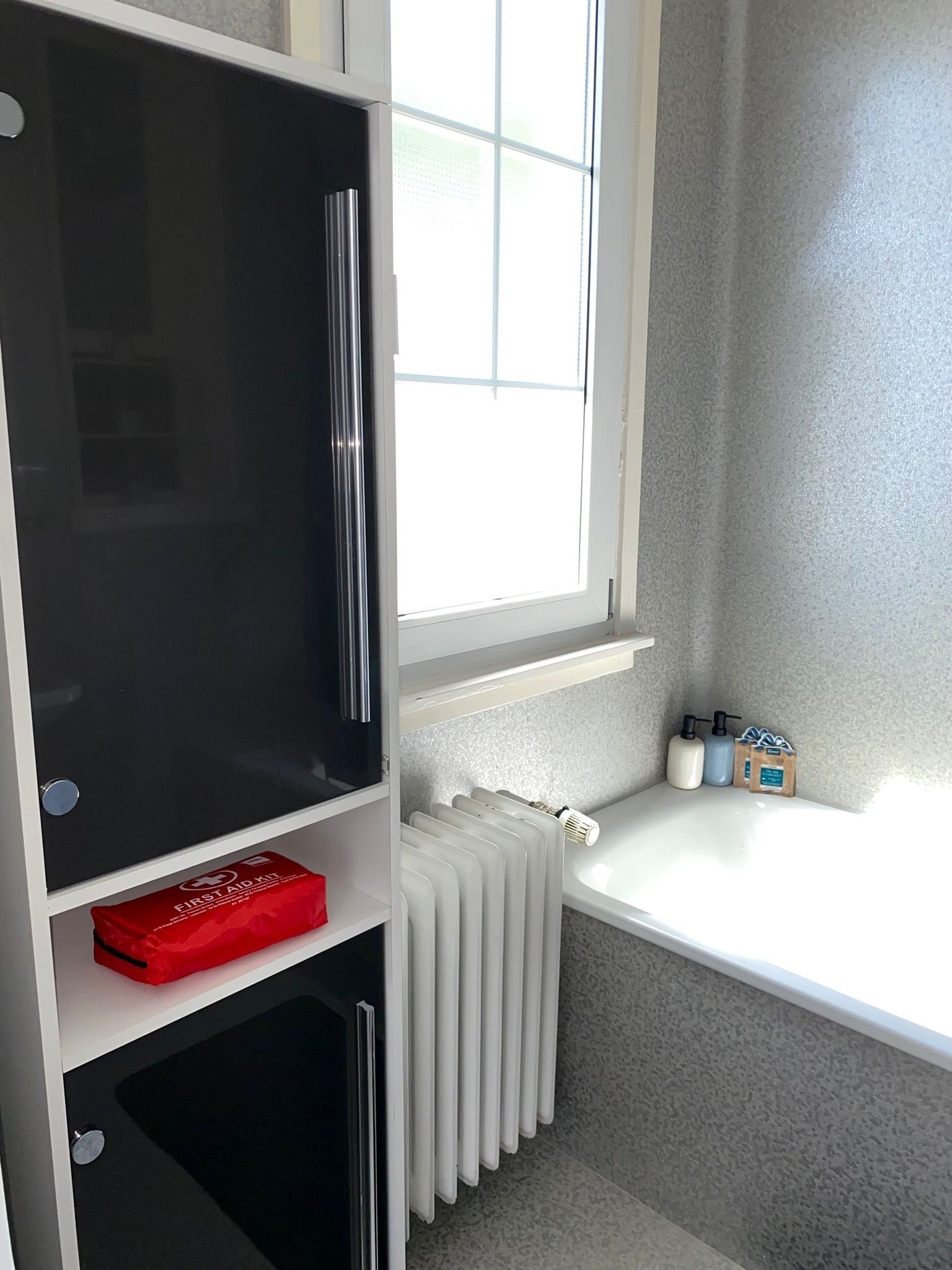 Una habitación con ventana, un radiador y un estante con una caja roja que dice "Botiquín de Primeros Auxilios".