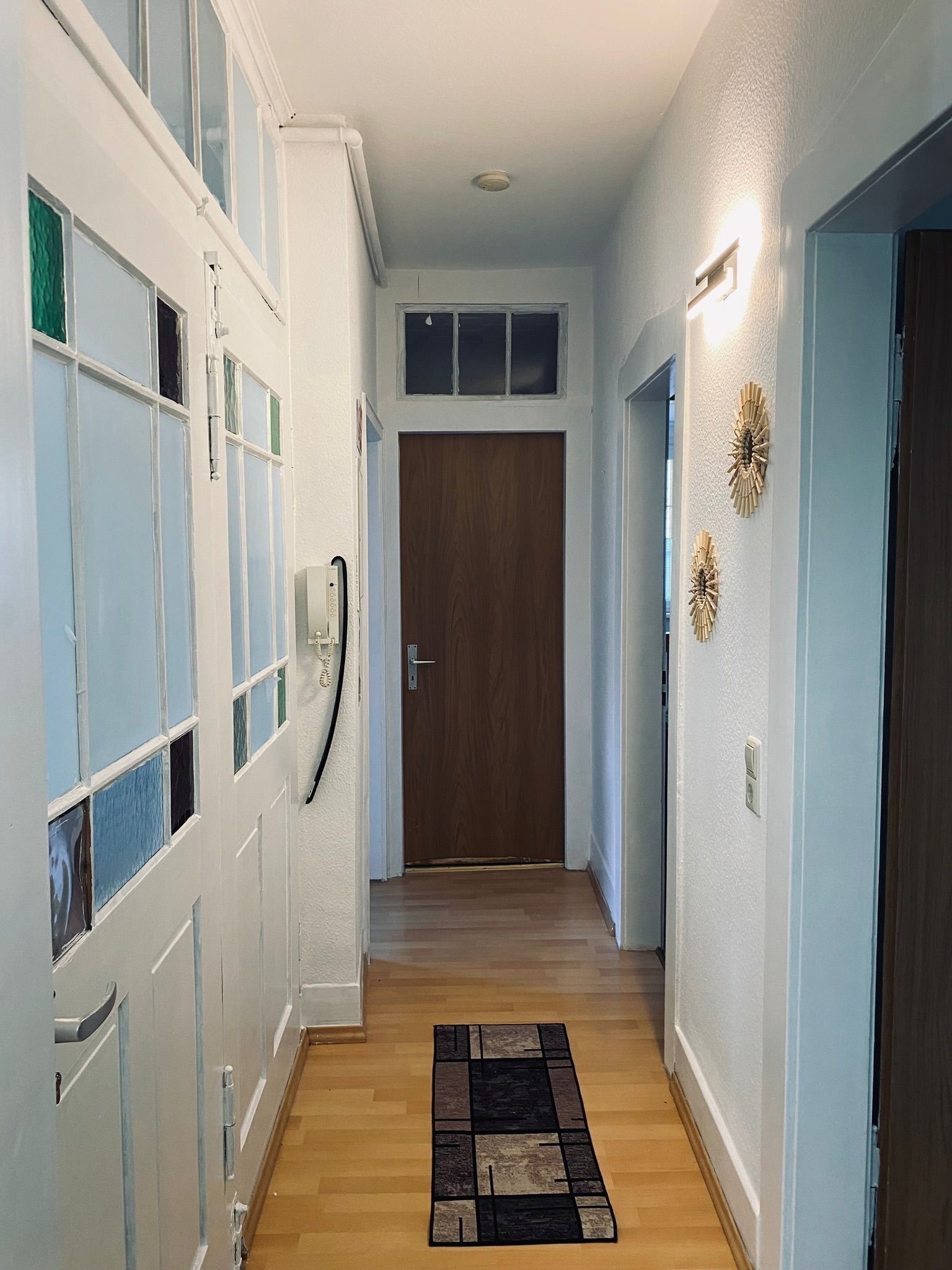 A hallway with a wooden door at the end, a light switch on the wall, and a decorative wall piece.