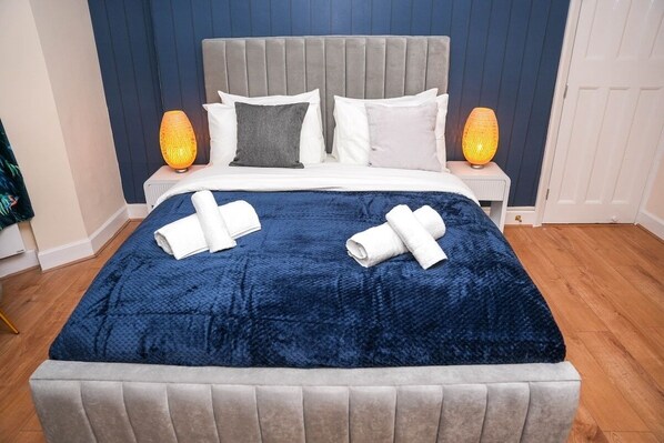 Ein ordentlich bezogenes Bett mit einer blauen Decke, zwei Kissen und zwei gerollten Handtüchern.