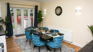 Un coin repas avec une table dressée pour quatre personnes, des chaises à l’assise rembourrée de couleur bleue, une horloge au mur et, par une porte vitrée, une vue sur l’extérieur.