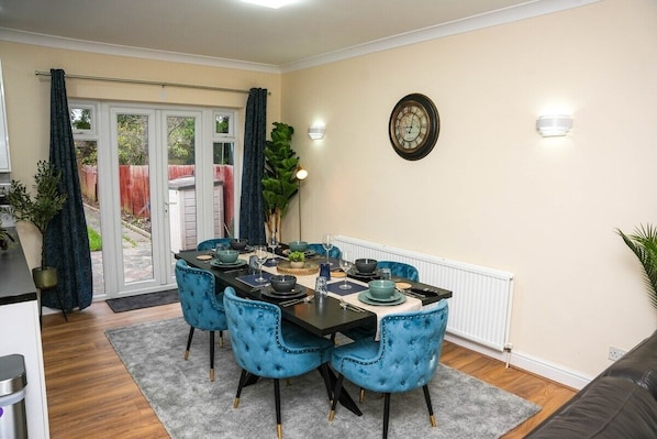 A dining area with a table set for four, blue upholstered chairs, a clock on the wall, and a view through a glass door to the outside. - Spacious 4-Bed Contractor Home | Heathrow & M4 | Free Parking (Hounslow)